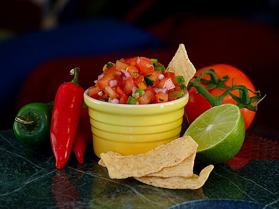 Fresh salsa in a yellow bowl surrounded by colorful ingredients and tortilla chips.