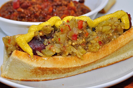 Toasted hot dog with relish and mustard, served alongside a bowl of hearty chili.