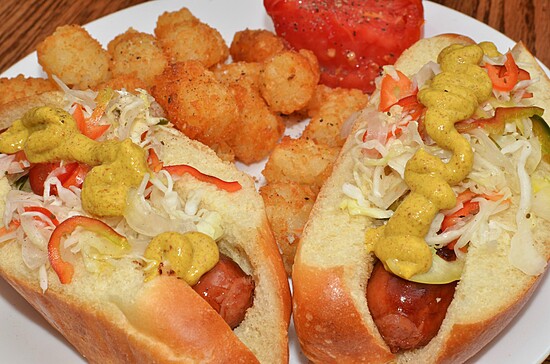 Juicy hot dogs with mustard, crunchy toppings, and crispy tater tots on a white plate.